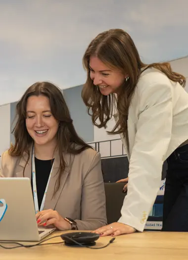 Twee jonge vrouwen in een kantoorruimte, lachend achter een laptop met het FrieslandCampina-logo en de naam Maud op een sticker; op de achtergrond een fotowand van een distributiecentrum.